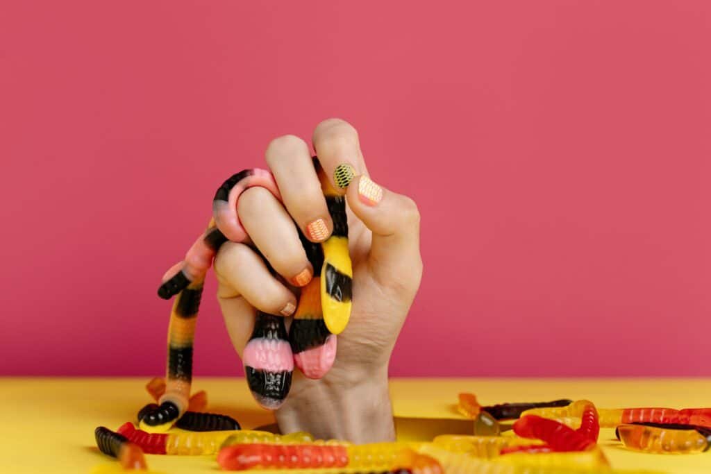 Hand gripping colorful gummy worms with bright nail art, set against a pink and yellow backdrop.

