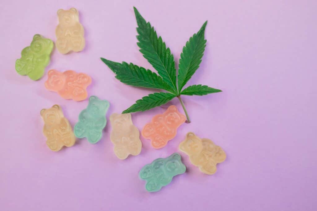 Colorful gummy bears and a cannabis leaf arranged on a pastel purple background. 