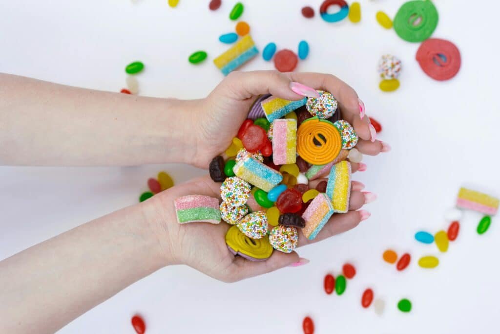 Hands full of bright, assorted candy gummies over a white background with scattered pieces.

