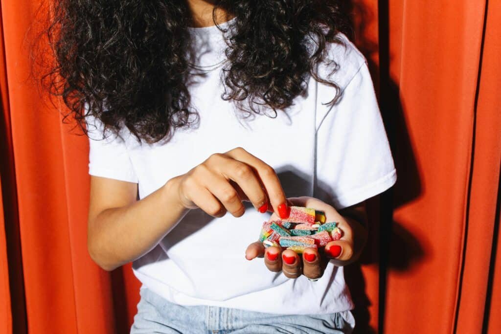 Person with curly hair holding colorful sour gummies in their hands, wearing a white t-shirt.