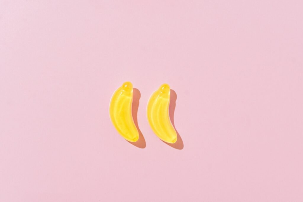 Two banana-shaped yellow gummies on a pink background, shown in bright natural lighting. 