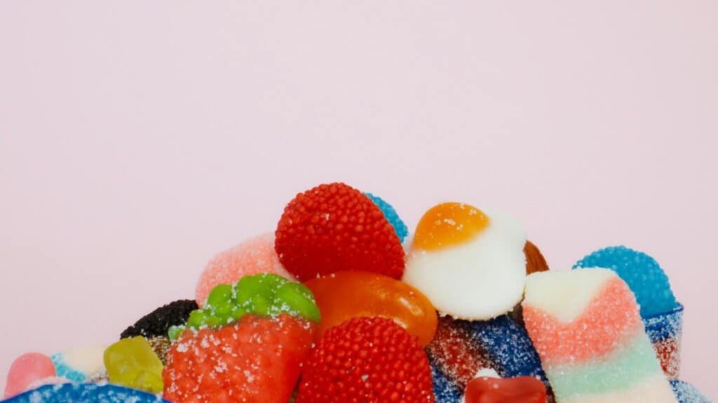Assorted sugar-coated gummy candies including strawberries, eggs, and rainbow strips on a pastel pink background.

